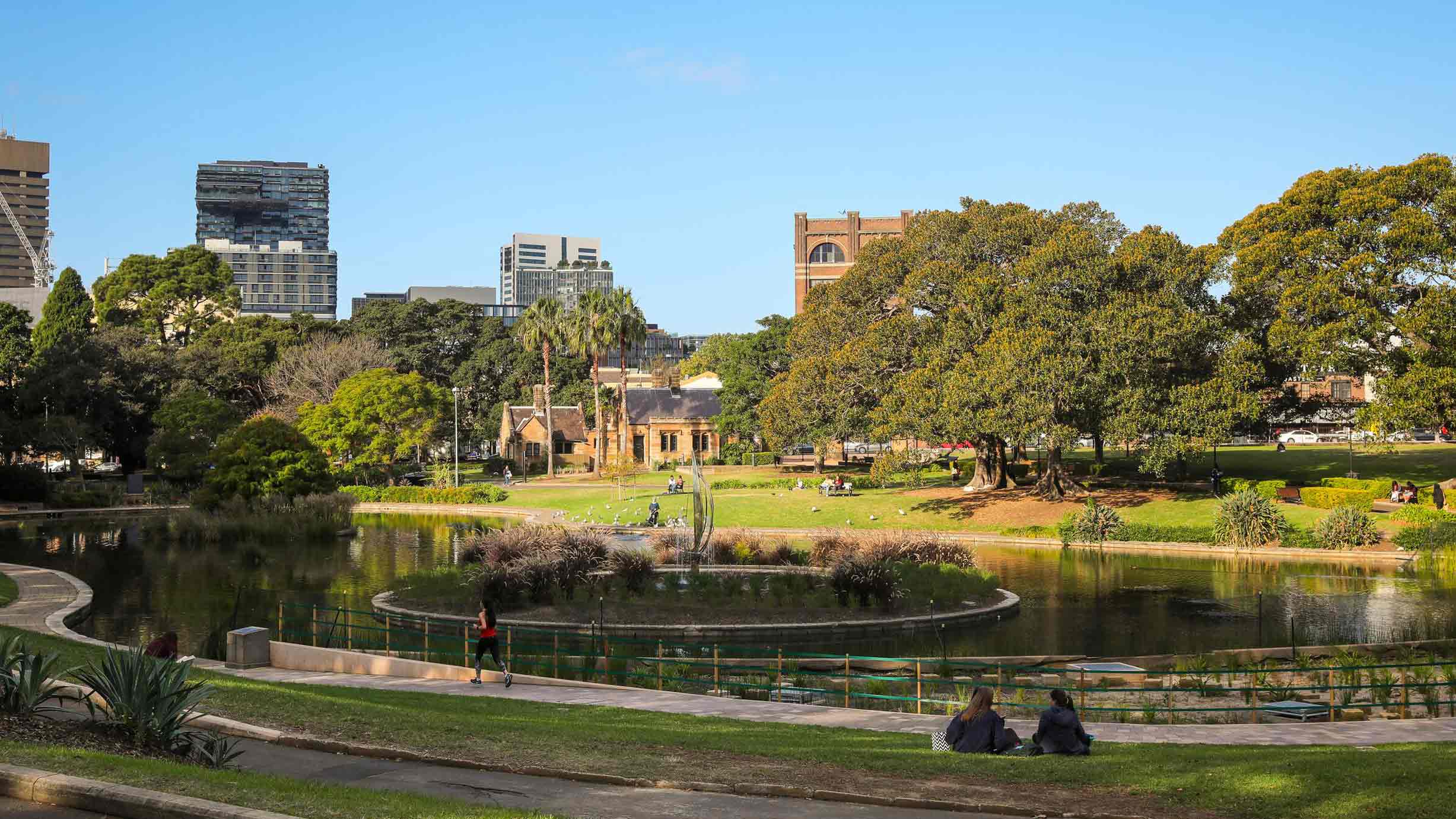 A city park with a pond, central fountain, trees, and people sitting on the grass; buildings are visible in the background under a clear sky.